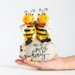 Close-up of two yellow Smadeer honey bee figurines holding flowers, sitting on a stone with an inscription.